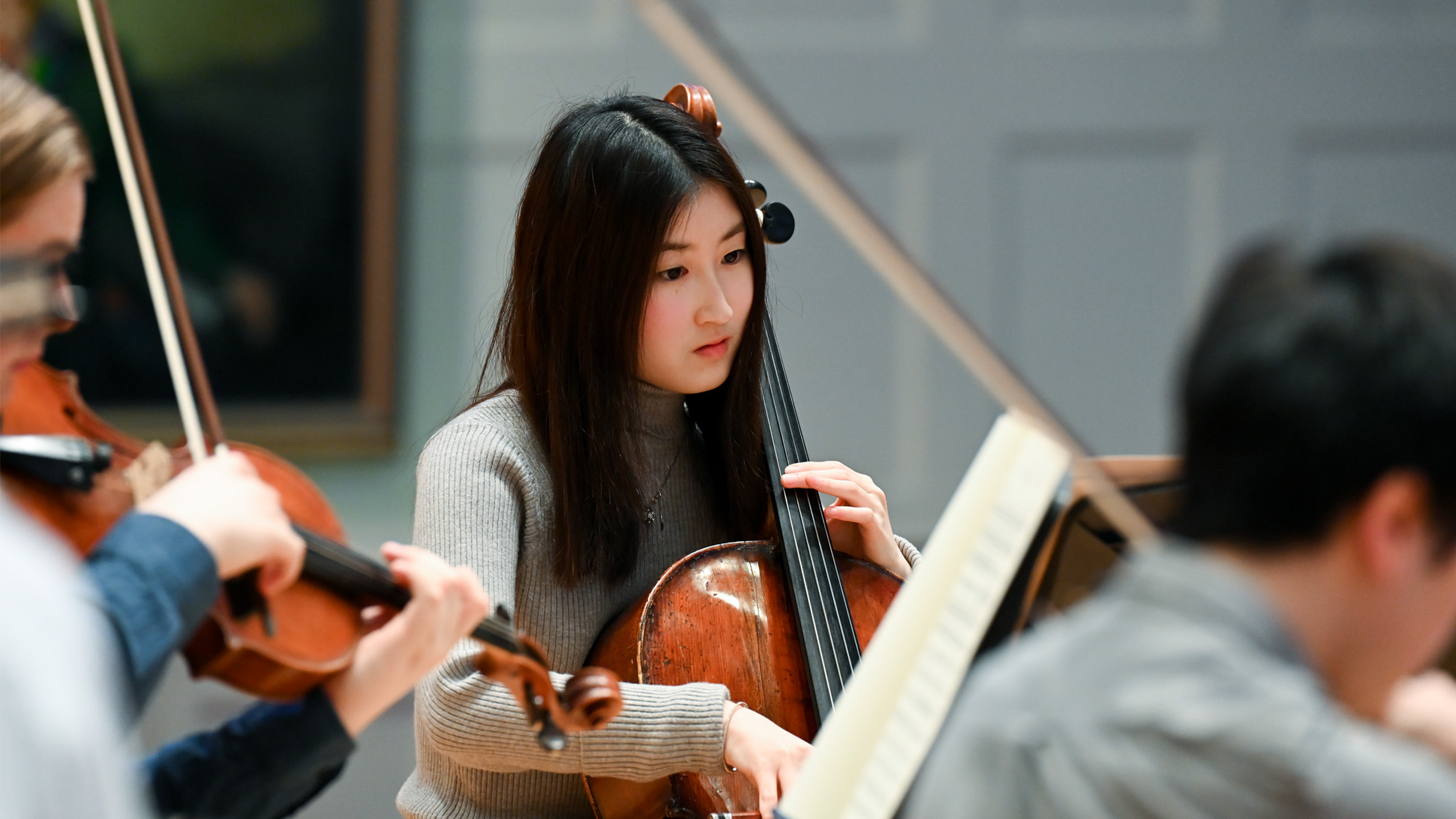 An Asian student, focusing on playing the cello in an orchestra rehearsal.
